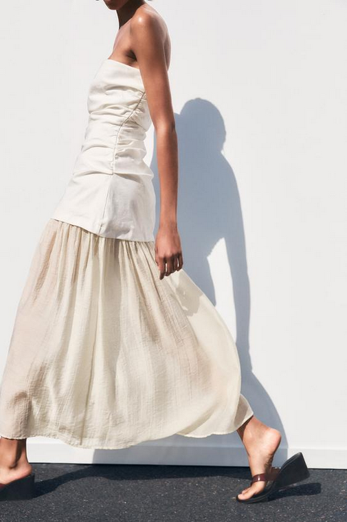 Woman wearing a white strapless top and beige skirt against a white wall.