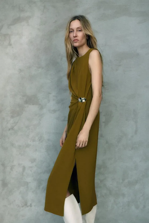 Woman wearing a long olive green dress with a belt against a gray background